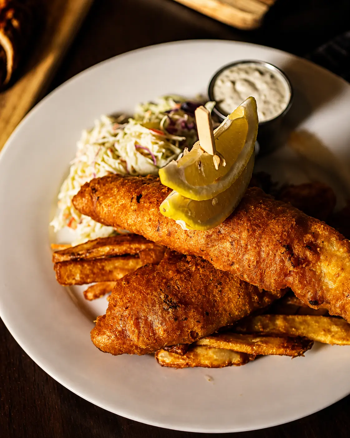 Two golden beer-battered fish fillets over thick-cut chips with coleslaw, tartar sauce and a lemon wedge on a white plate.