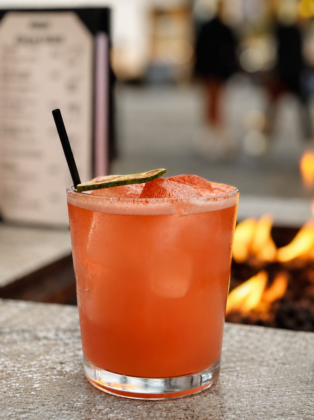 The Spicy Flamingo cocktail, a chili-rimmed grapefruit drink with a dehydrated lime wheel, on the patio bar beside a lit fire table.