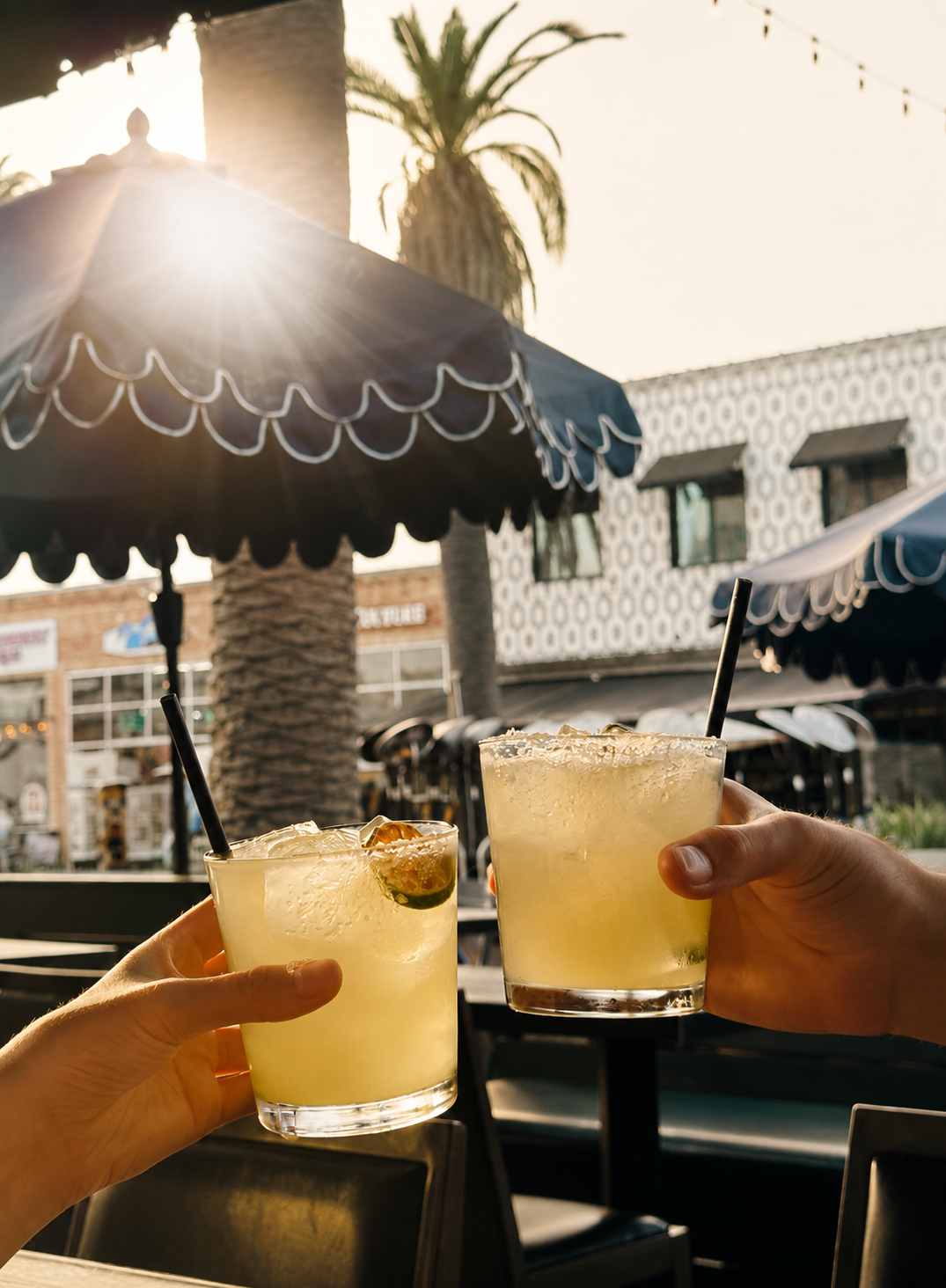 Two guests clinking tall cocktails over a patio table at golden hour, sunlight flaring behind a navy scalloped umbrella and palm trees on Pier Avenue.